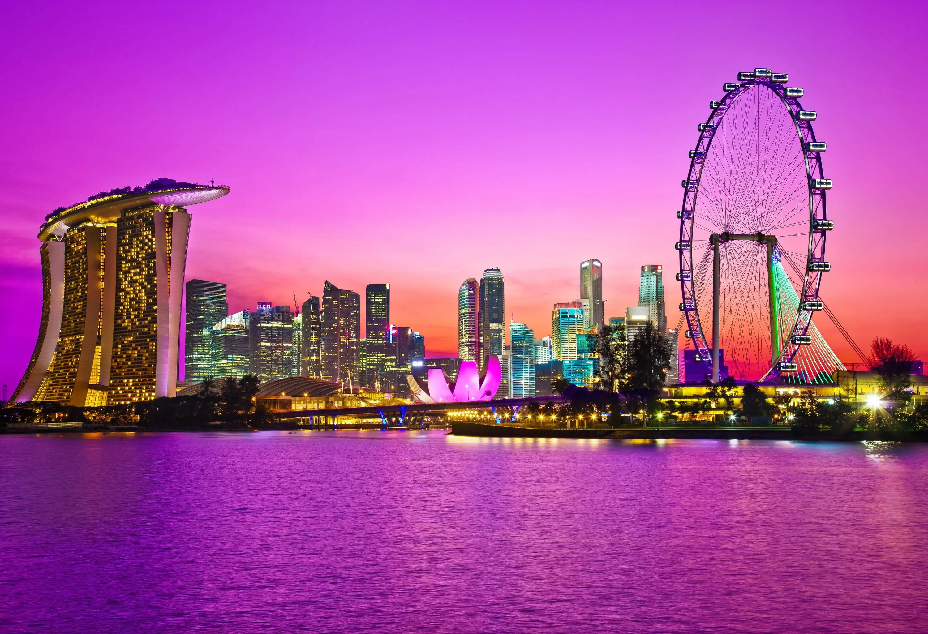 During dusk, the breathtaking Singapore landmark city skyline at Marina Bay shines brightly with illuminated buildings, including the iconic Marina Bay Sands and the futuristic ArtScience Museum, while the nearby Singapore Flyer adds an enchanting touch as it stands tall by the coast.