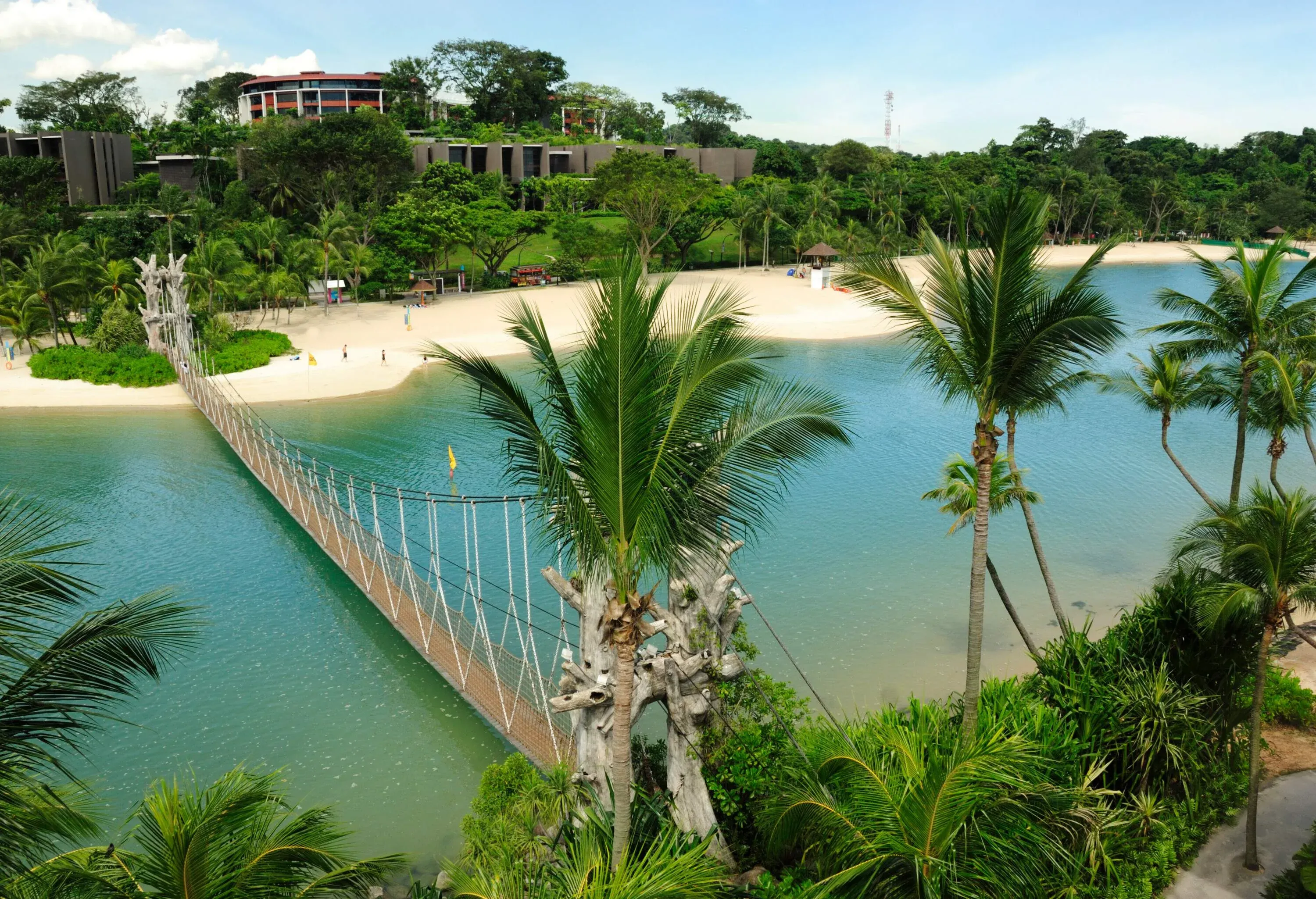 A rope bridge over the turquoise sea connects two lush islands with a white sandy shore and modern beach houses.